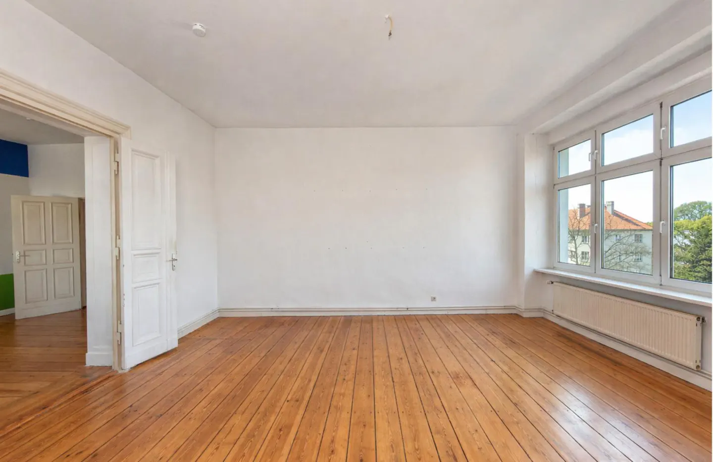 Bright, empty room with hardwood floors, white walls, and a large window showing a building and trees. A white door is slightly ajar.