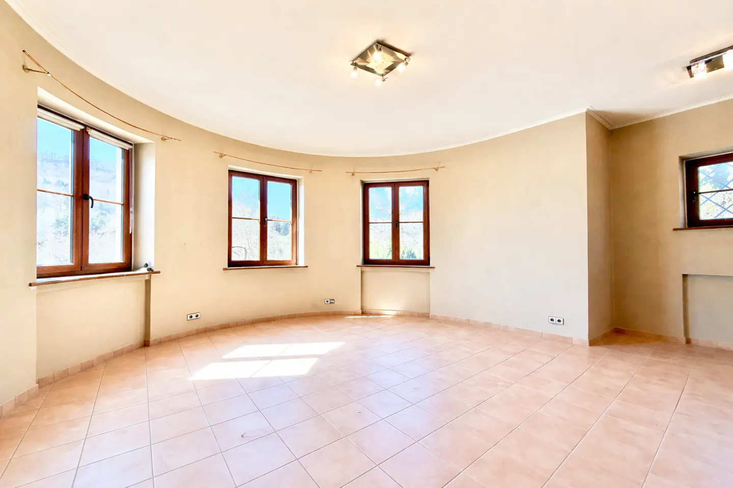 Empty room with curved walls, beige paint, and brown-framed windows. The floor is tiled in a diamond pattern.