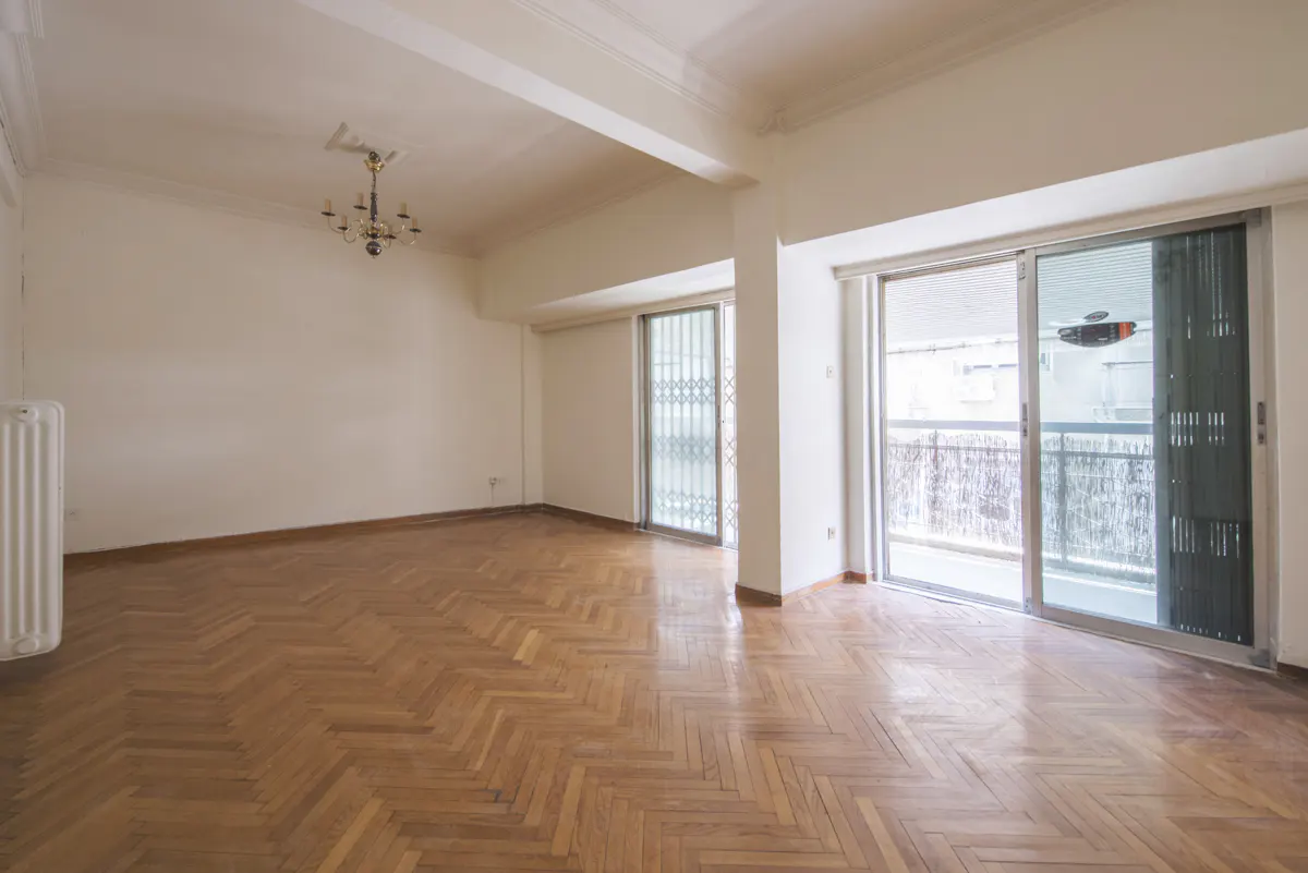 Empty room with light walls, herringbone wood floors, a vintage chandelier, and sliding glass doors leading to a balcony.