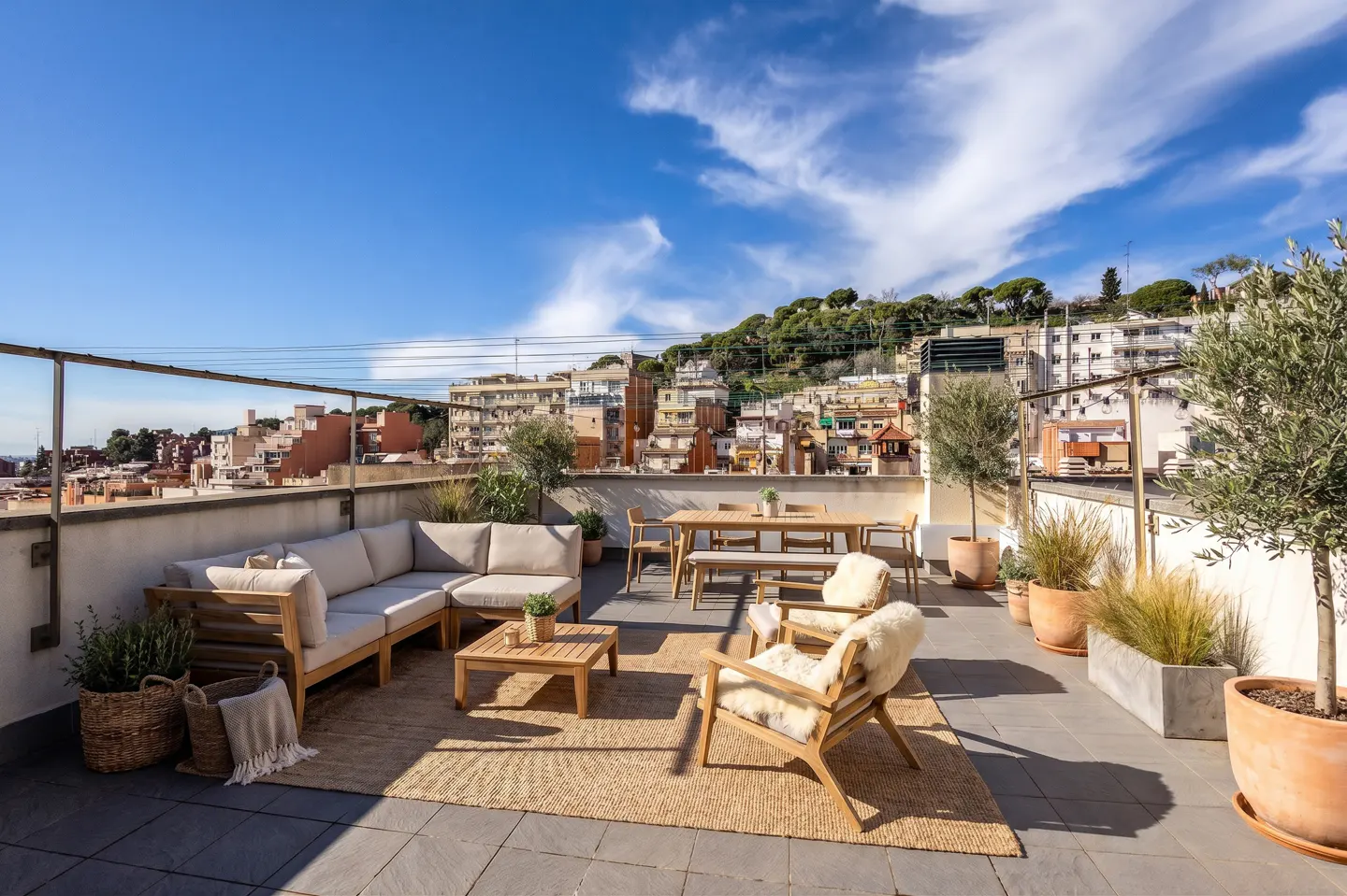 Sunny rooftop terrace with wooden furniture, a beige sectional sofa, dining set, and potted plants against a city backdrop.