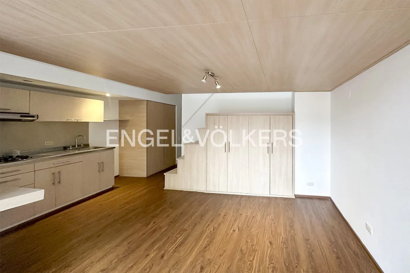 A bright, open-concept apartment with wood floors, a kitchen with beige cabinets, and stairs with built-in storage.