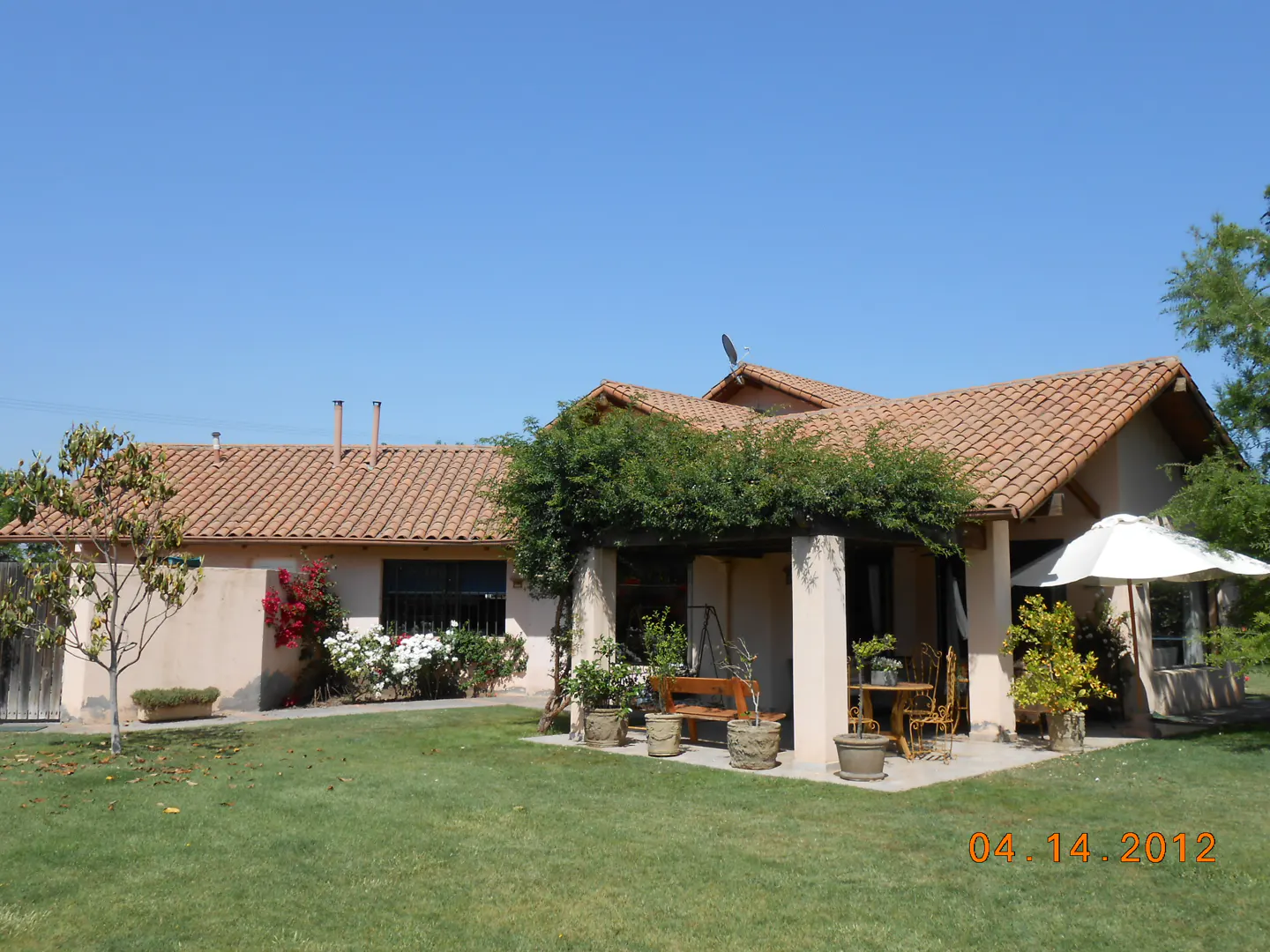 Exterior view of a tan house with a red tile roof, green lawn, and patio with furniture.
