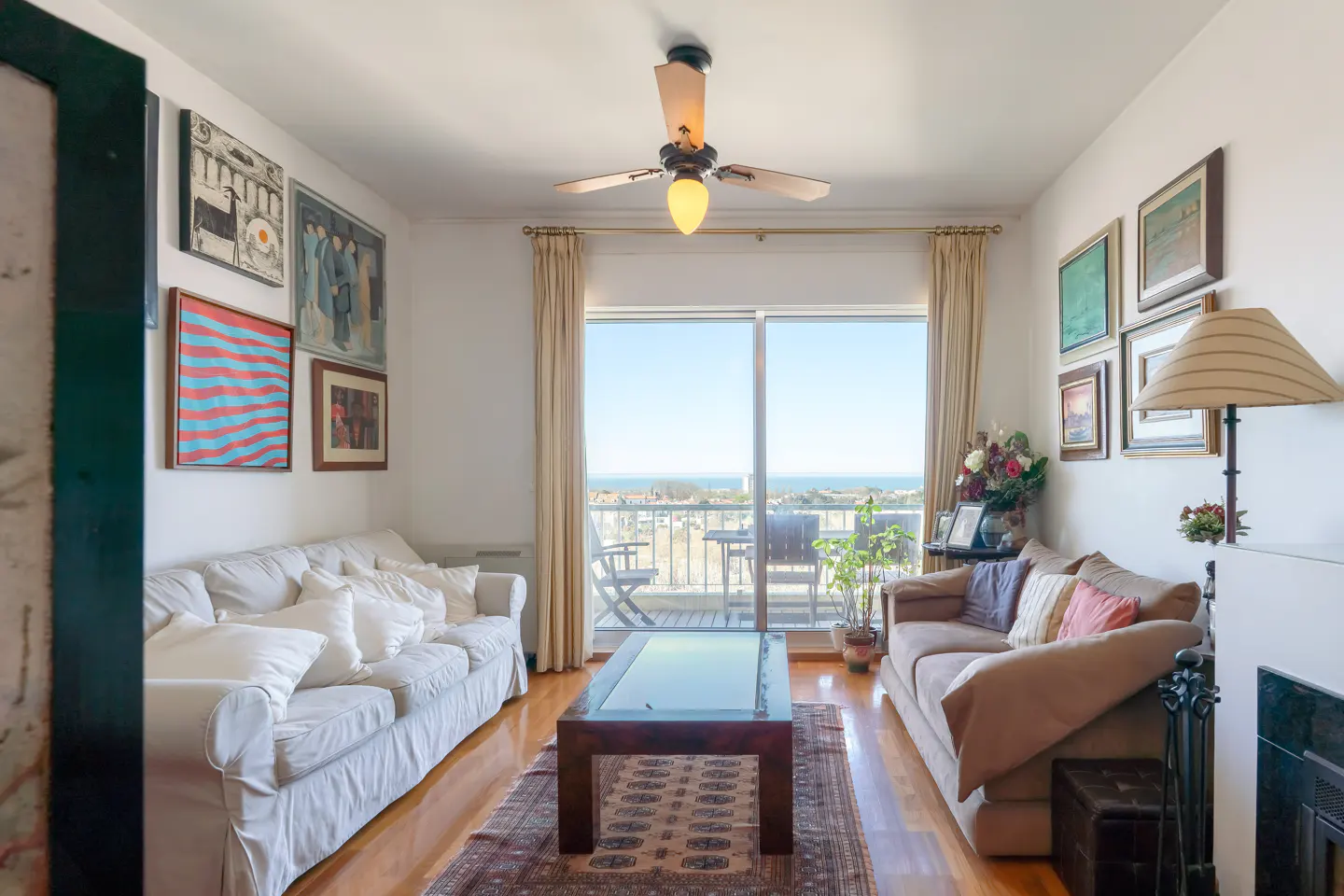 Living room with white and brown sofas, wood floors, and a glass coffee table. Balcony with ocean view.