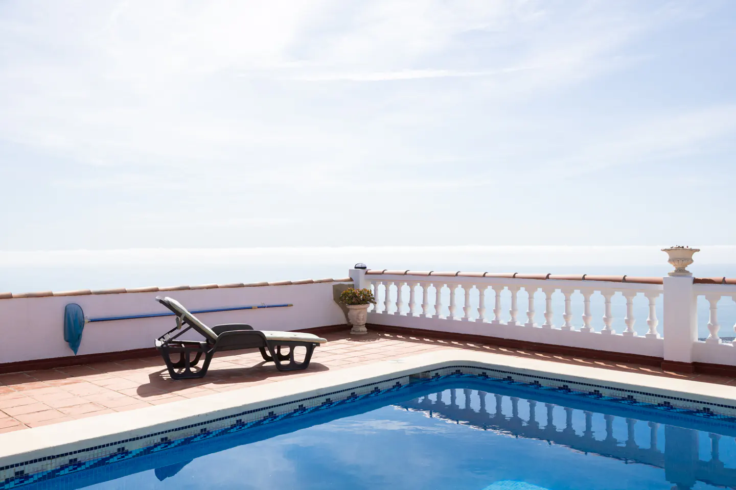 Outdoor pool with blue water reflecting the sky, a lounge chair, and a white balustrade overlooking the ocean.