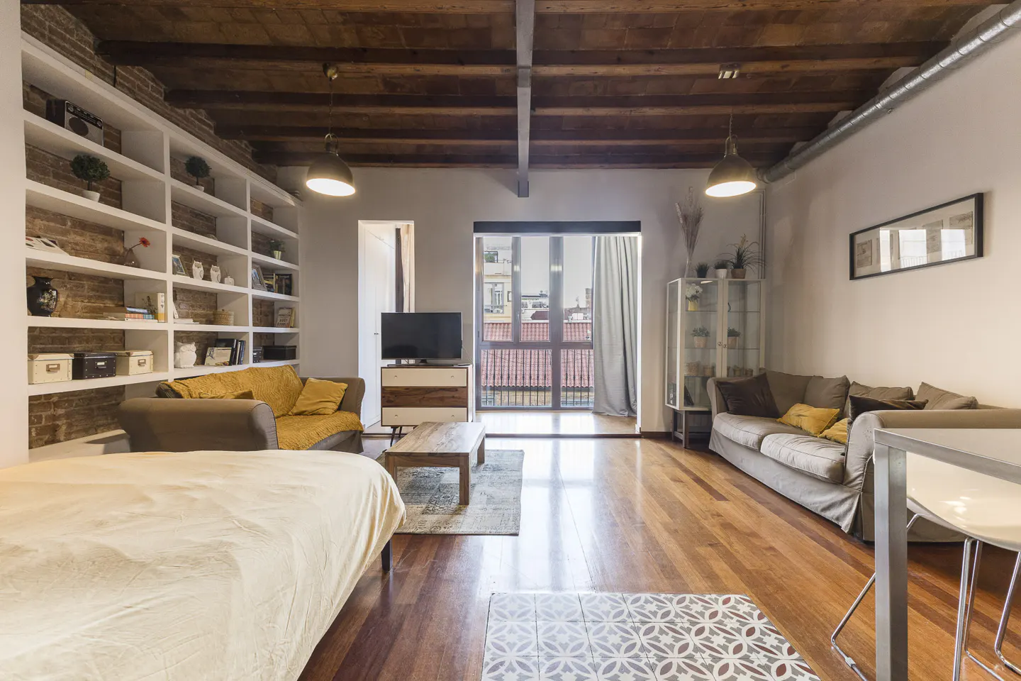 Bright living room with hardwood floors, exposed wood beam ceiling, and a wall of built-in bookshelves. A bed, sofas, and a balcony are also visible.