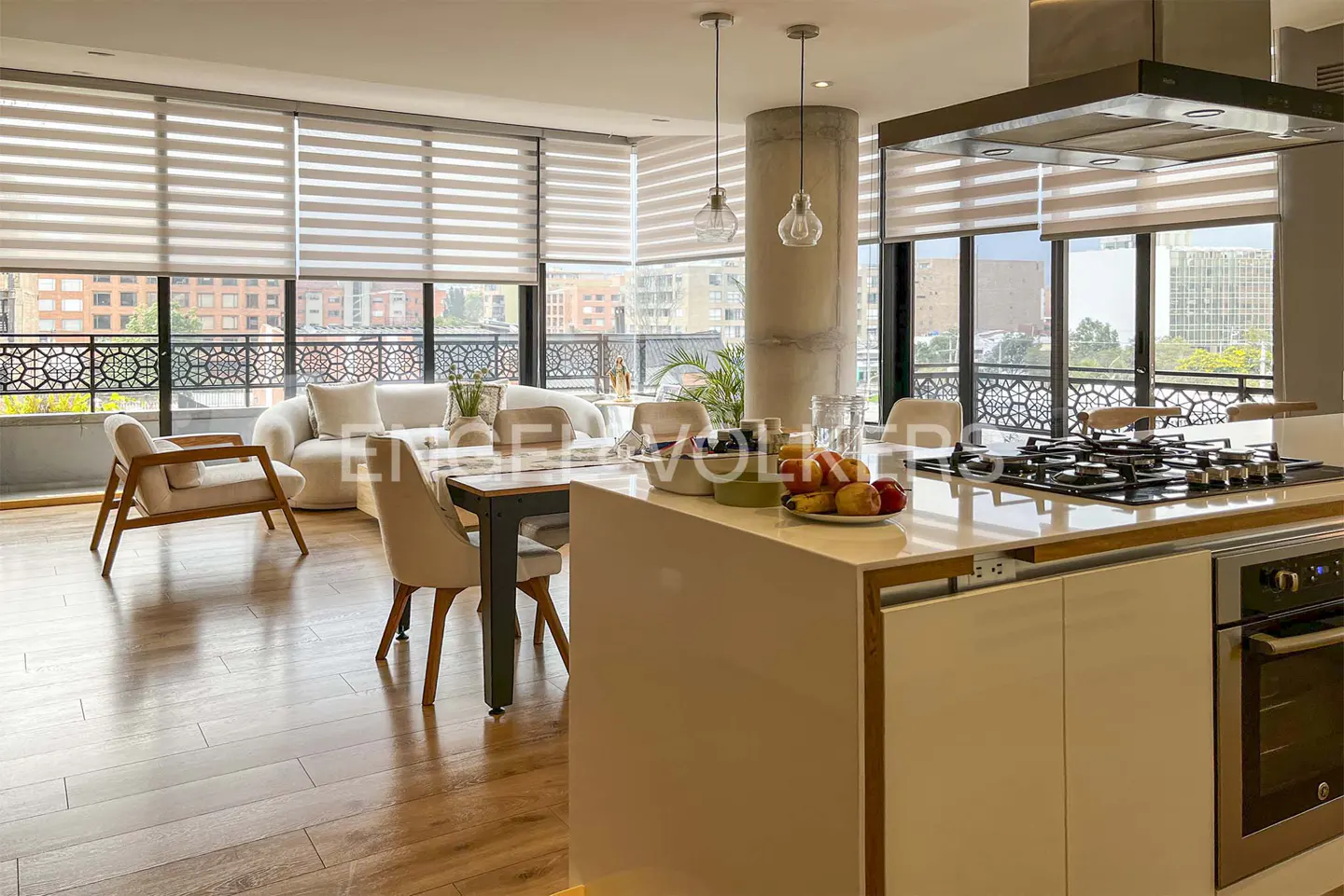Open-concept living space with wood floors, a white sofa, dining table, and kitchen island with a gas cooktop. Large windows offer city views.