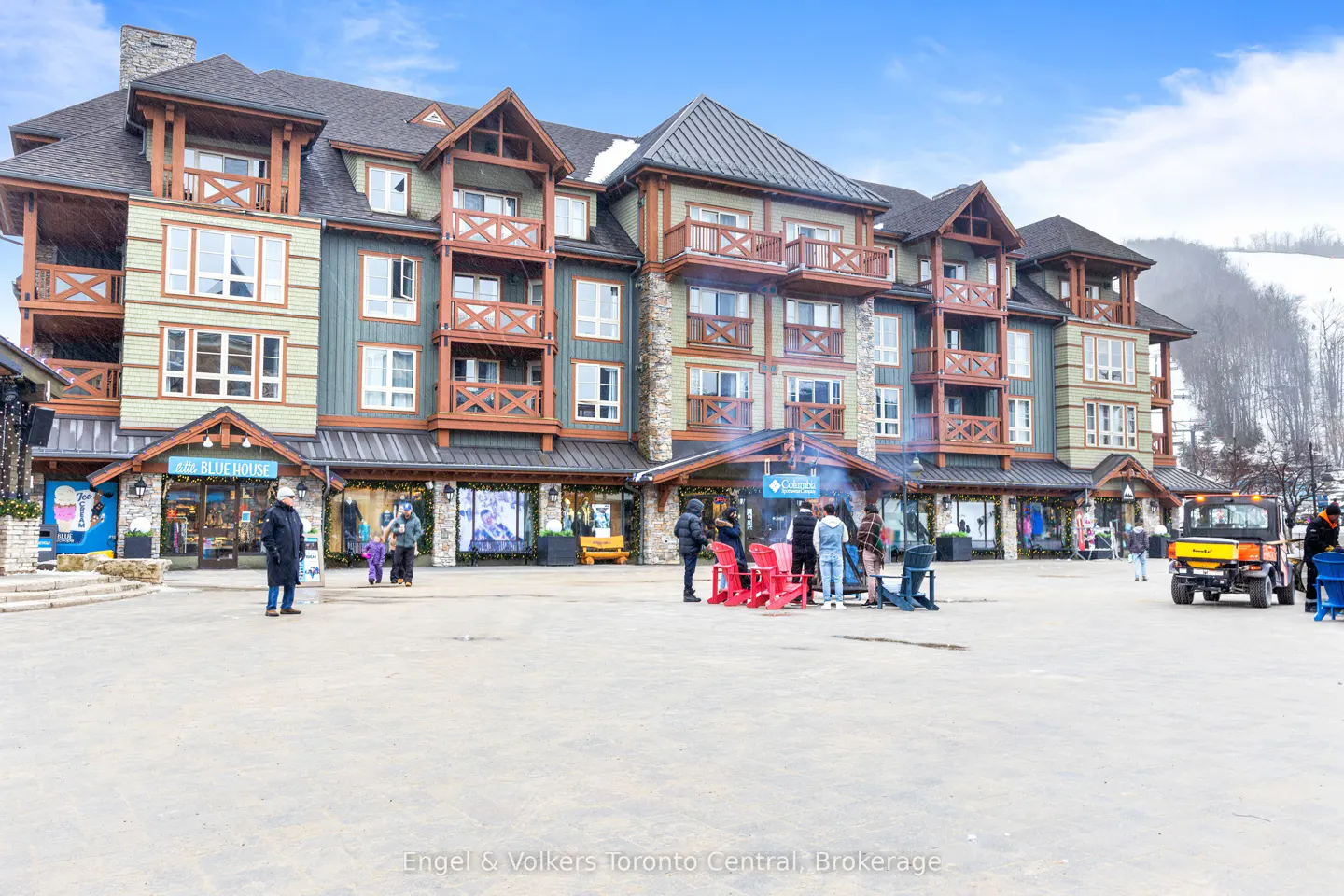 Multi-story condo building with shops on the ground floor. People gather around a fire pit with red chairs. Ski hill in the background.