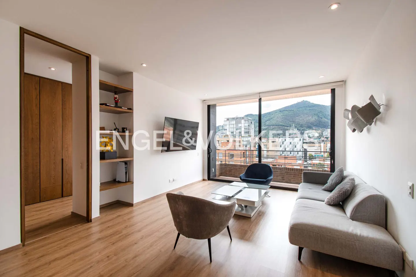 Bright living room with wood floors, gray sofa, blue chair, and city view through large windows. A TV hangs on the wall.