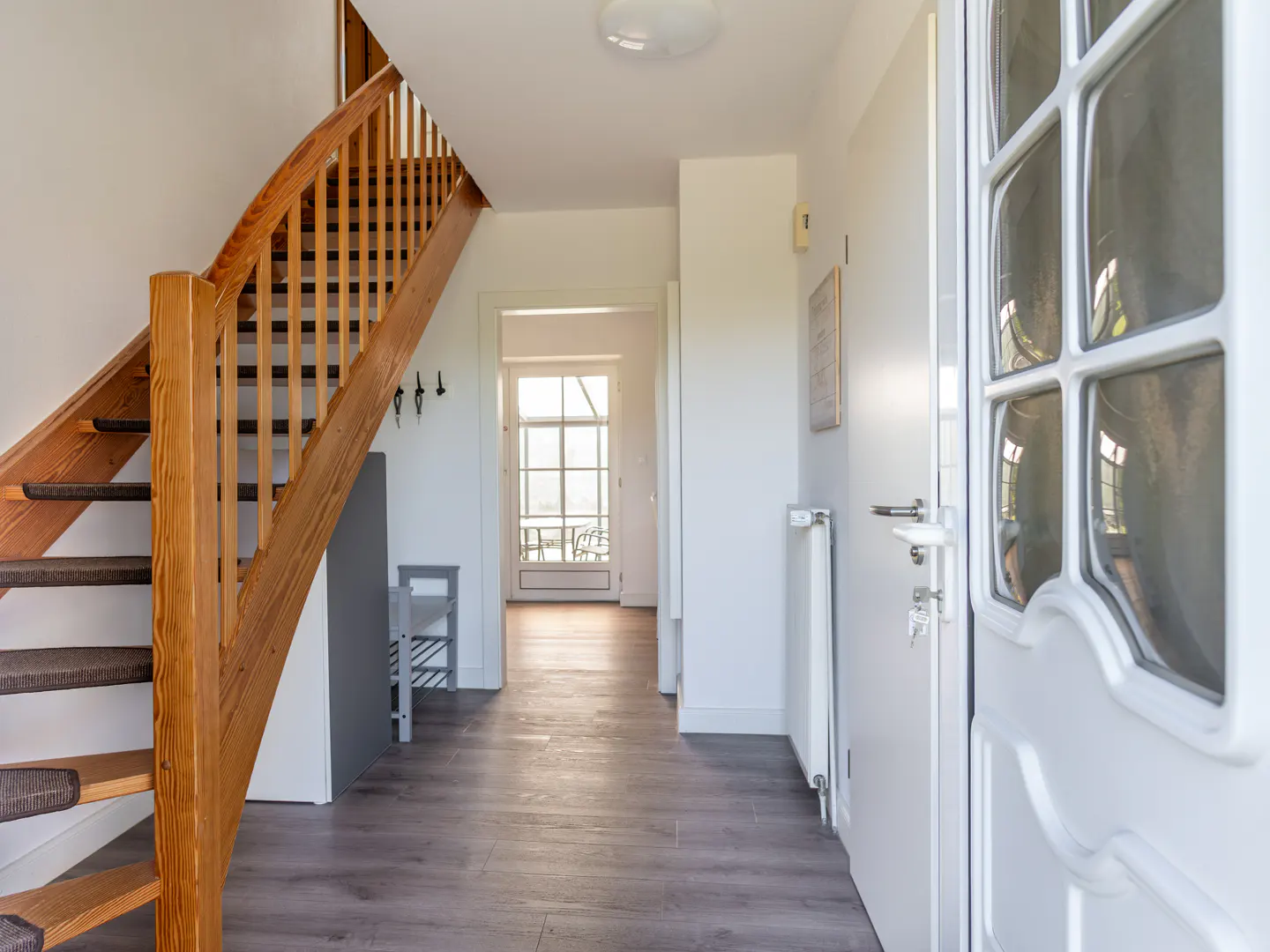 Bright entryway with a wooden staircase, grey wood-look flooring, and a white door leading to a sunlit room.