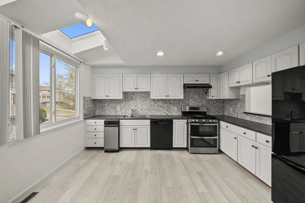 Bright kitchen with white cabinets, black appliances, and light wood floors. A large window and skylight provide natural light.