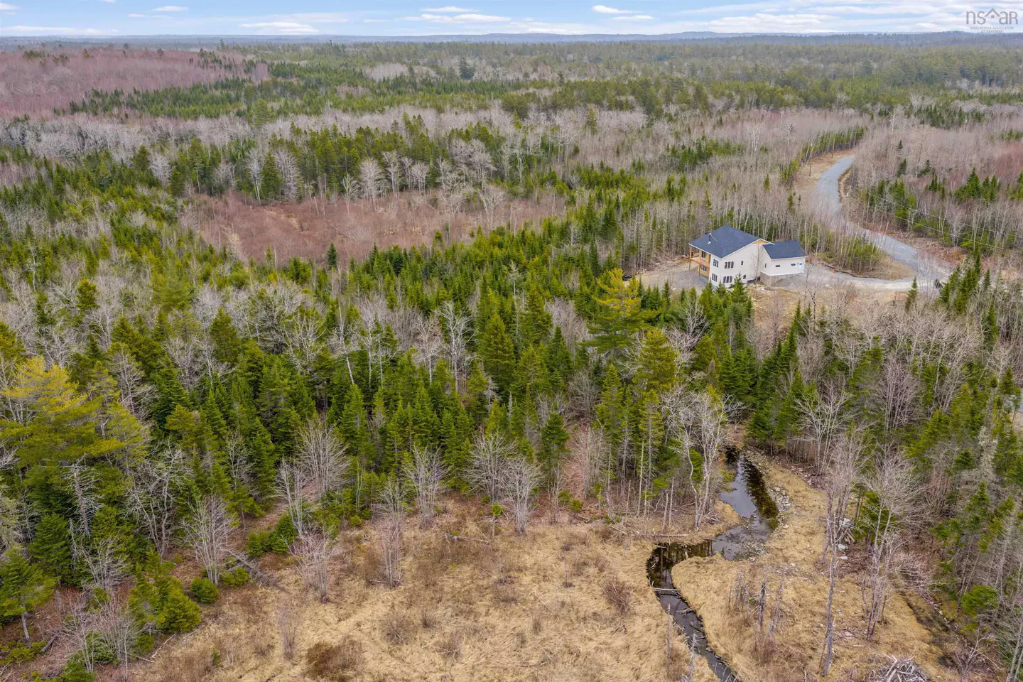 Aerial view of a light-colored house with a dark roof surrounded by a dense forest and a winding stream.