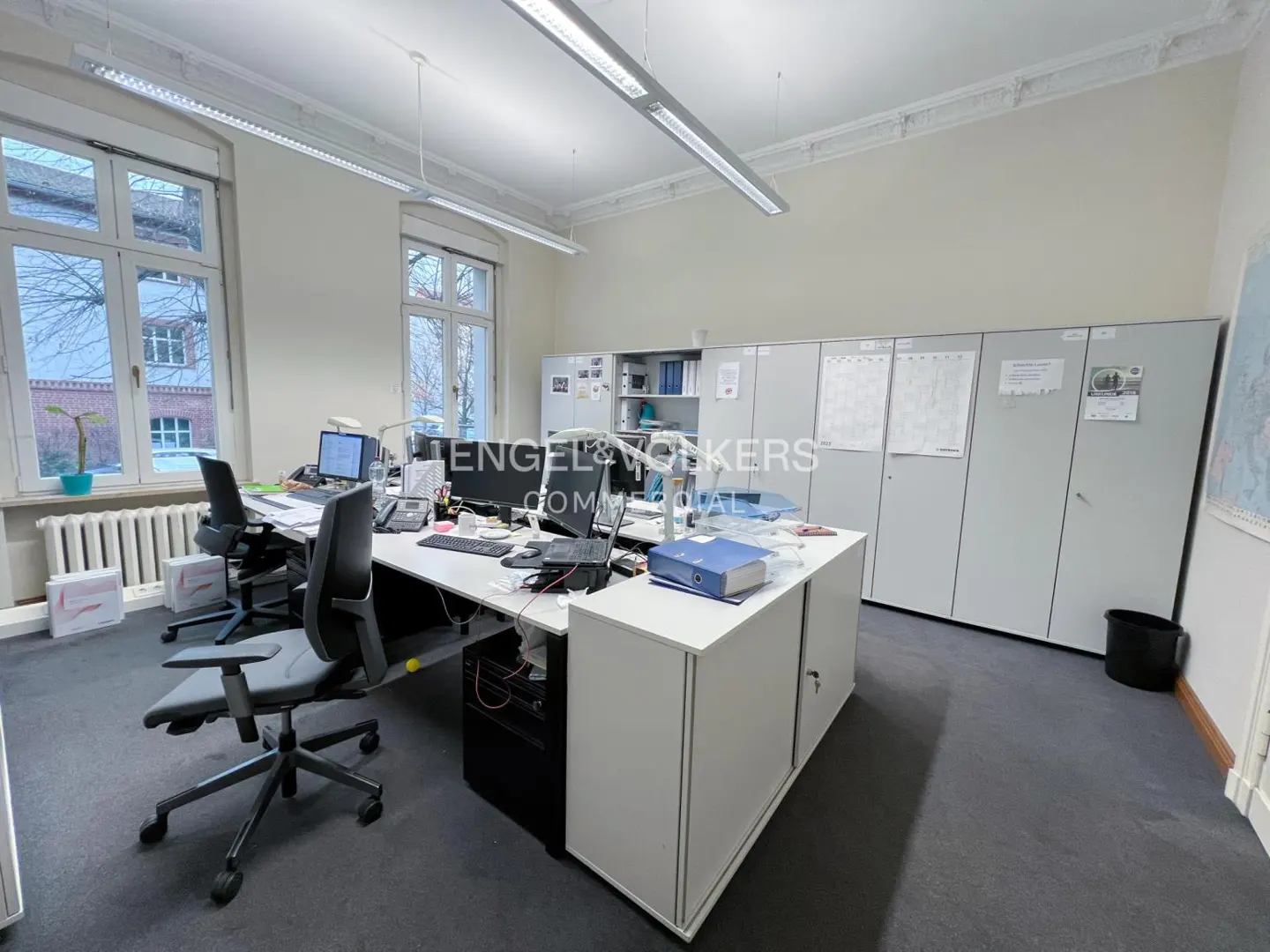 Bright office space with desks, chairs, computers, and storage cabinets. Large windows provide natural light. Gray carpet and white walls.