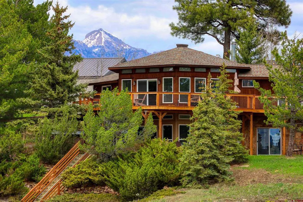 Two-story brown house with a deck, surrounded by green trees, with a snow-capped mountain in the background.