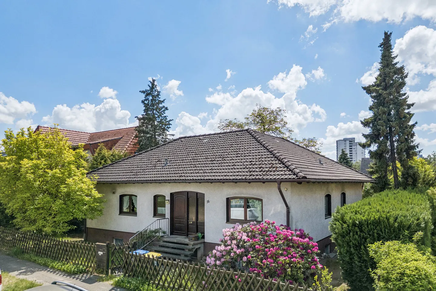 A one-story white house with a brown roof, surrounded by green trees and pink flowers, under a blue sky.