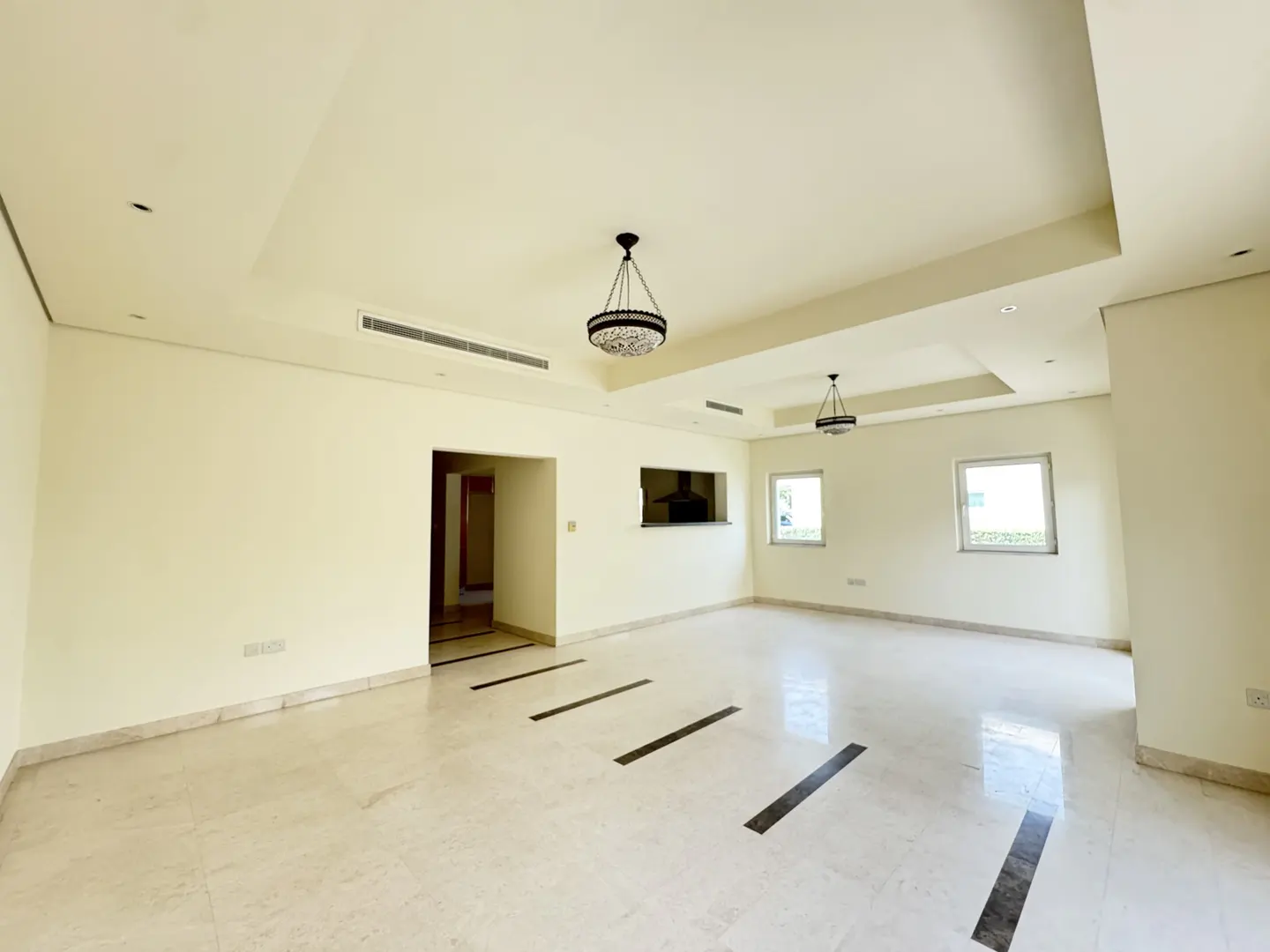 An empty, bright room with marble floors and cream walls. Two chandeliers hang from the ceiling. Two windows let in natural light.