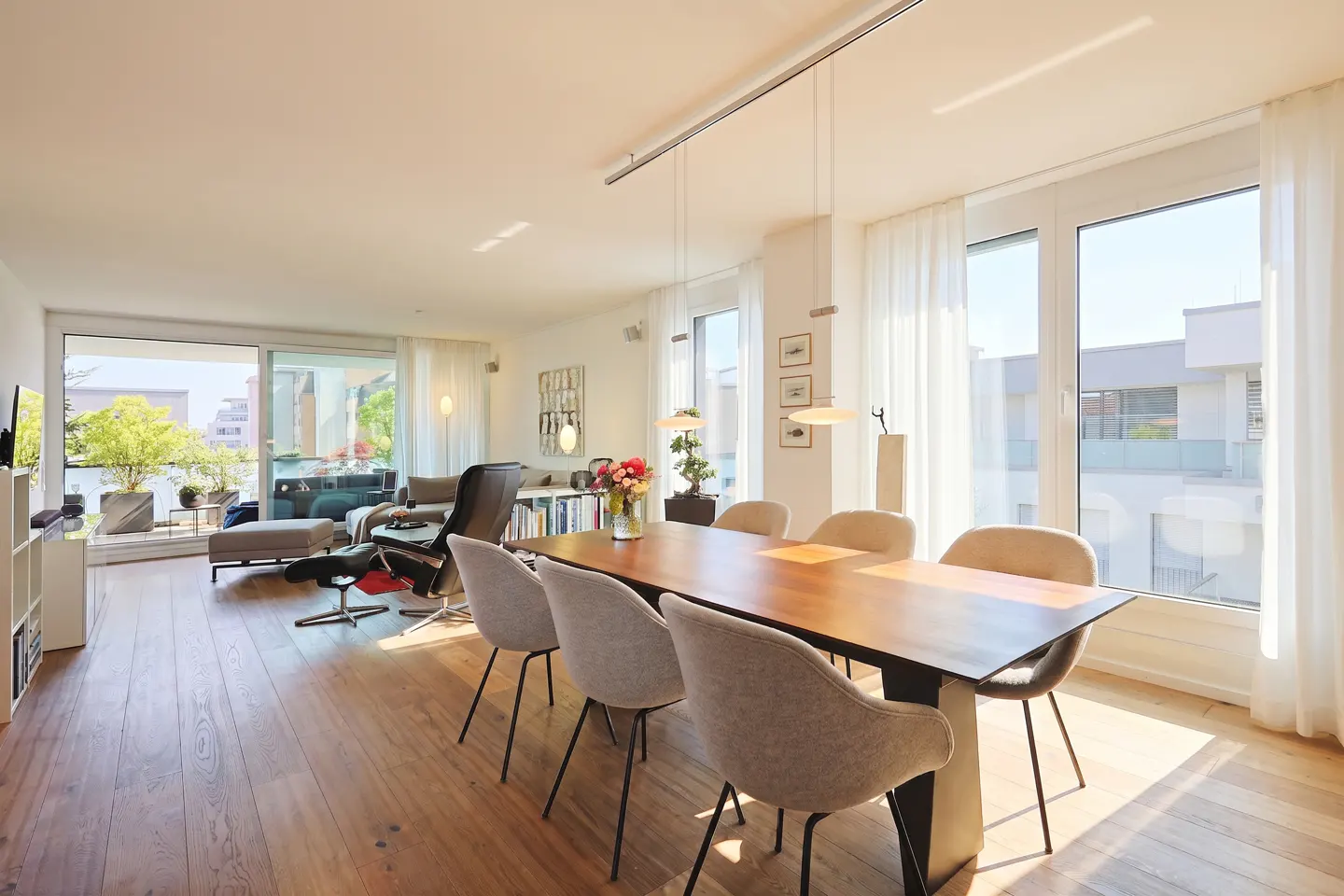 Bright, modern dining area with a wooden table, light gray chairs, and a living room with a balcony in the background.