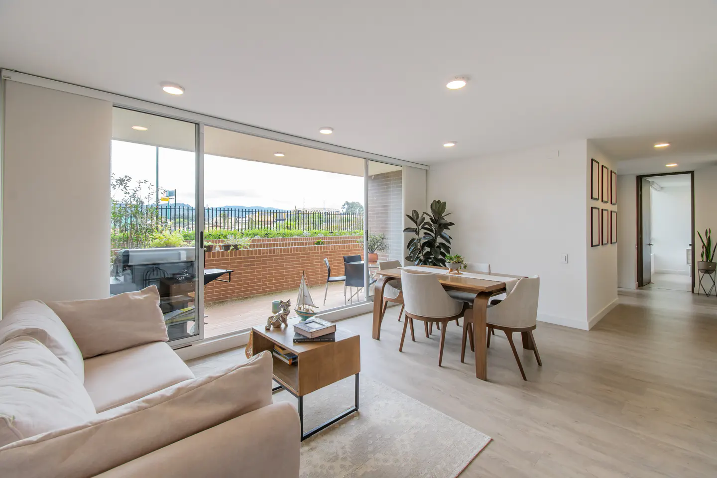 Bright living room with a beige sofa, wood table, and chairs. Sliding glass doors open to a balcony with a brick wall and green landscape.