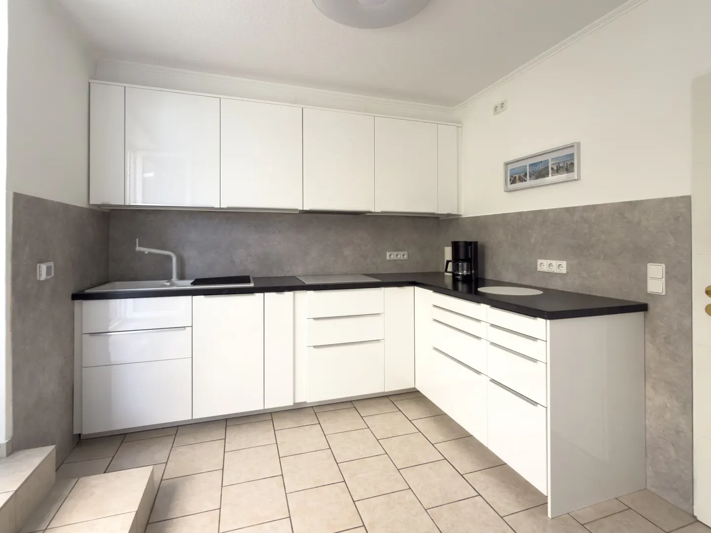 A modern kitchen with white cabinets, black countertops, and gray backsplash. Beige tile flooring. A coffee maker sits on the counter.