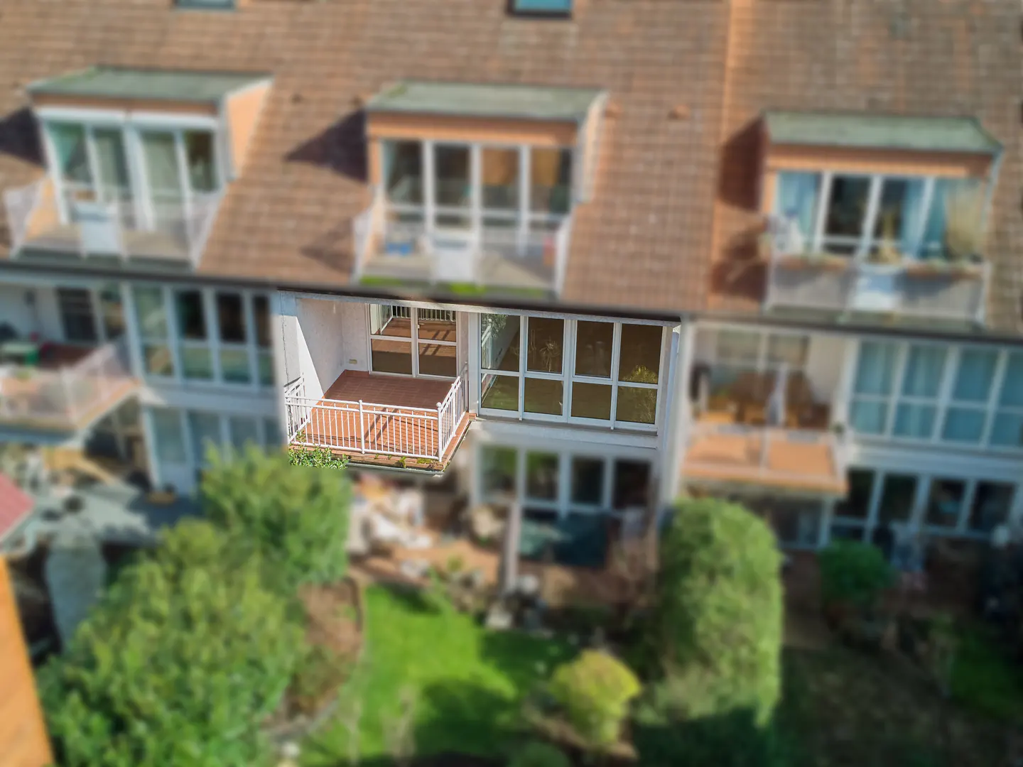 Aerial view of a townhouse with a brown roof, white trim, and a balcony overlooking a green garden.