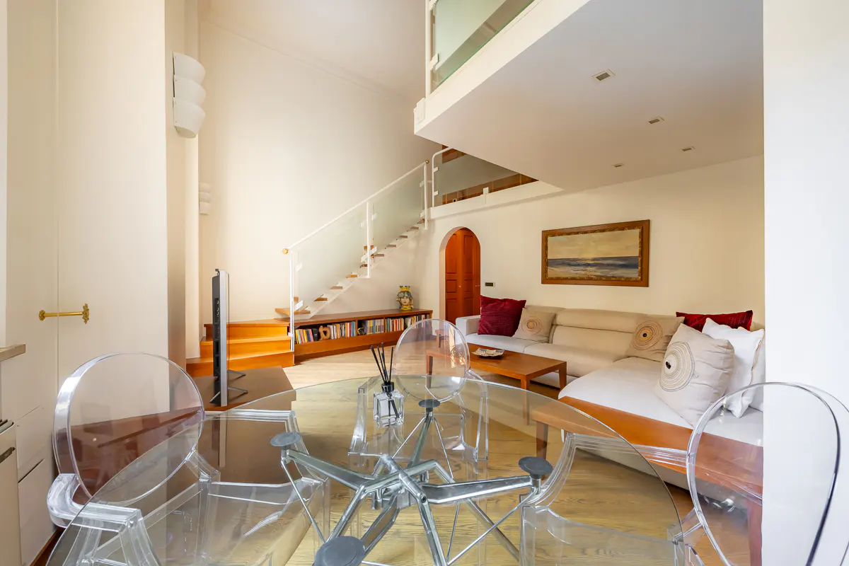 Bright, modern living room with a glass table and chairs in the foreground. A white sofa, stairs, and a painting are in the background.