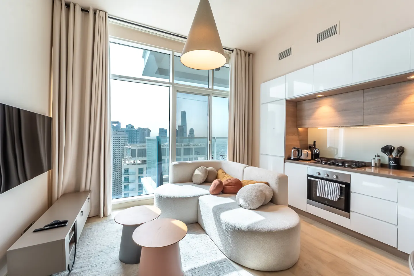 Bright apartment interior with a curved white sofa, modern kitchen, and a large window overlooking a city skyline.