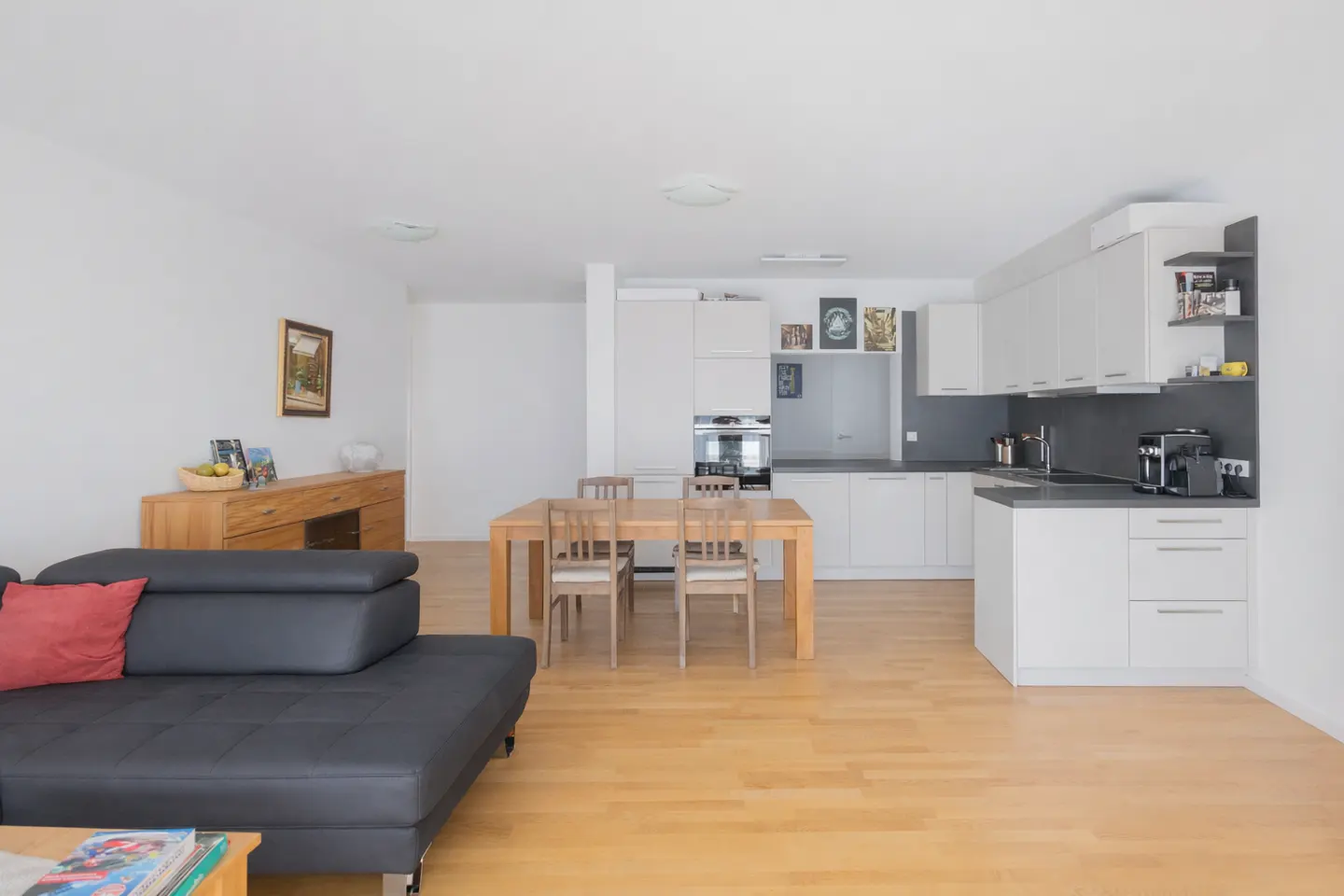Open-concept living space with a black sofa, wooden table and chairs, and a white kitchen with gray countertops.
