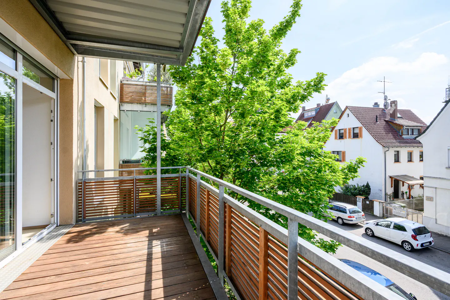 Balcony with wood floor and slatted railing overlooks a tree-lined street with houses and parked cars.