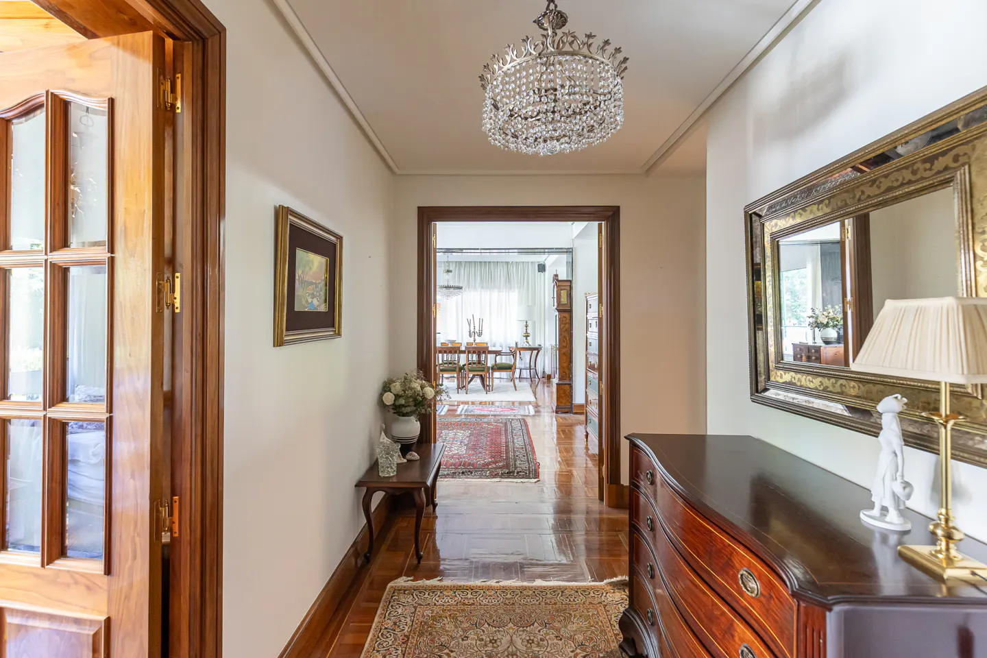 Elegant hallway with hardwood floors, crystal chandelier, wooden dresser, ornate mirror, and view into a dining room.