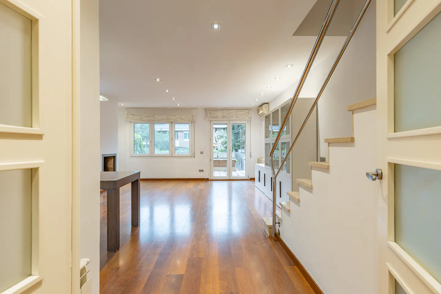 Open door view of a bright living room with hardwood floors, stairs with metal railing, and a table. Windows and recessed lighting illuminate the space.