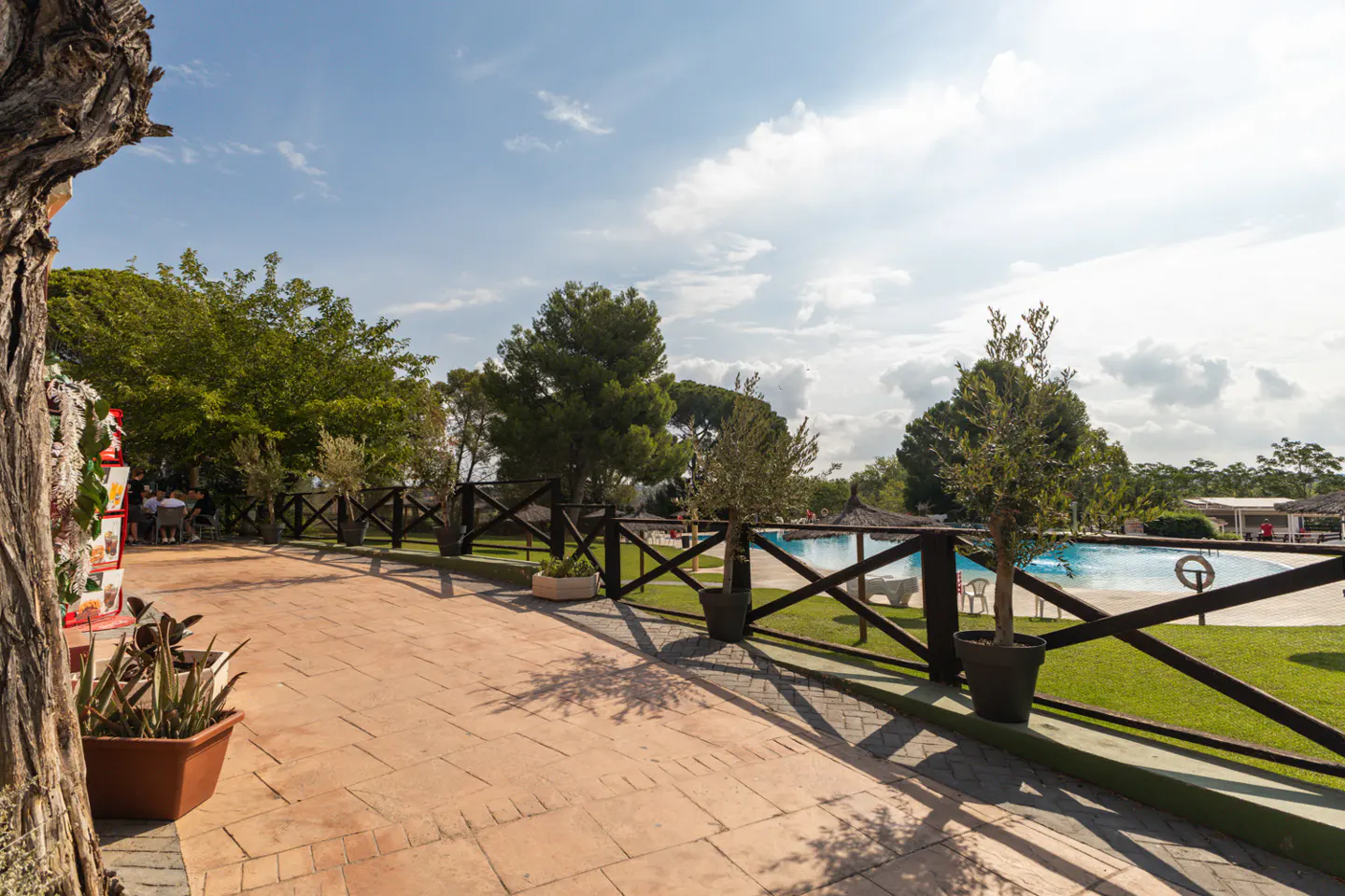 Outdoor patio with brick flooring and dark wood fence overlooking a pool area with green grass and trees under a blue sky.