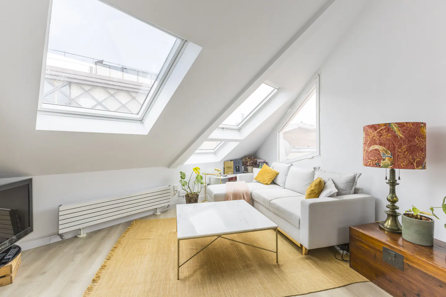 Bright attic living room with white walls, skylights, and a light gray sectional sofa with yellow pillows. A patterned lamp sits on a wooden chest.