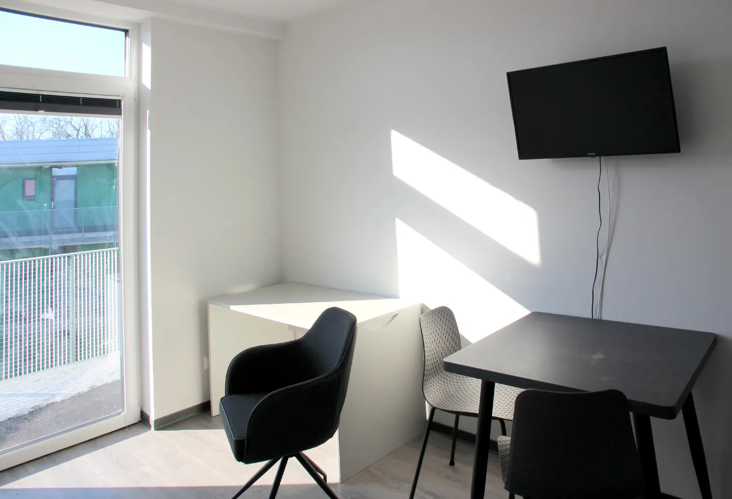 Bright room with a black table and chairs, a TV on the wall, and a window with a view of a building.