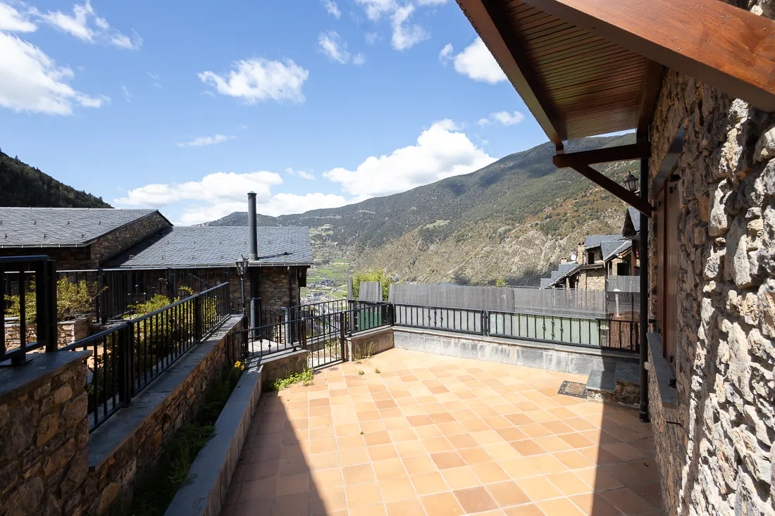 Stone house patio with terracotta tiles, black railings, and mountain views under a blue sky with clouds.