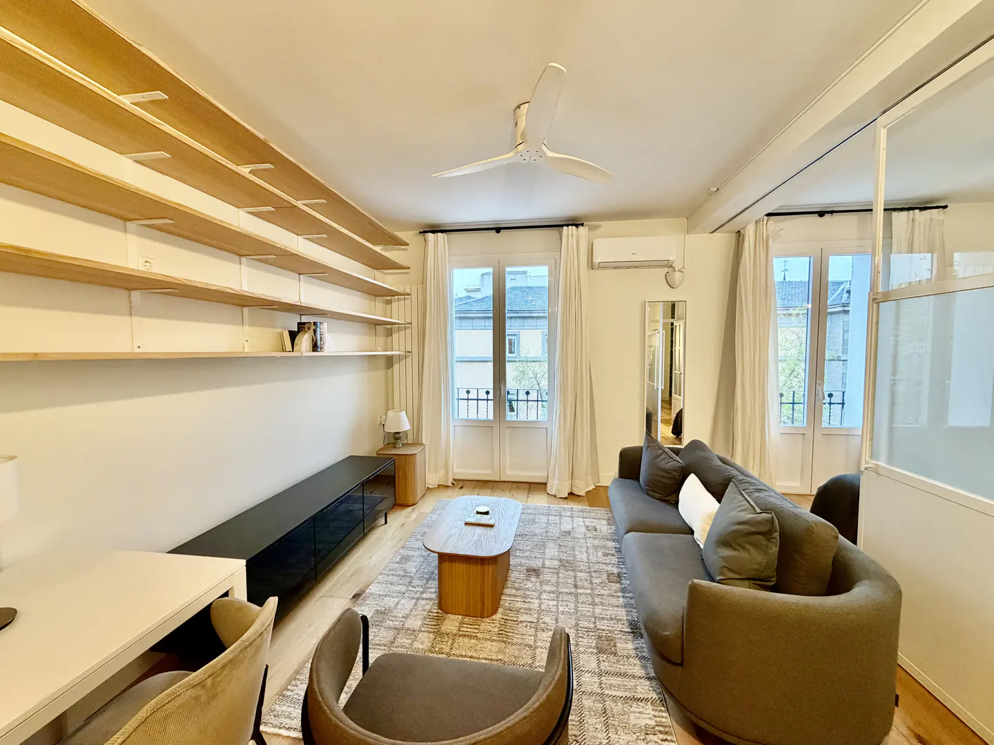 Bright living room with wood floors, a gray sofa, and a patterned rug. Open shelves line the wall above a black media console. French doors lead to a balcony.