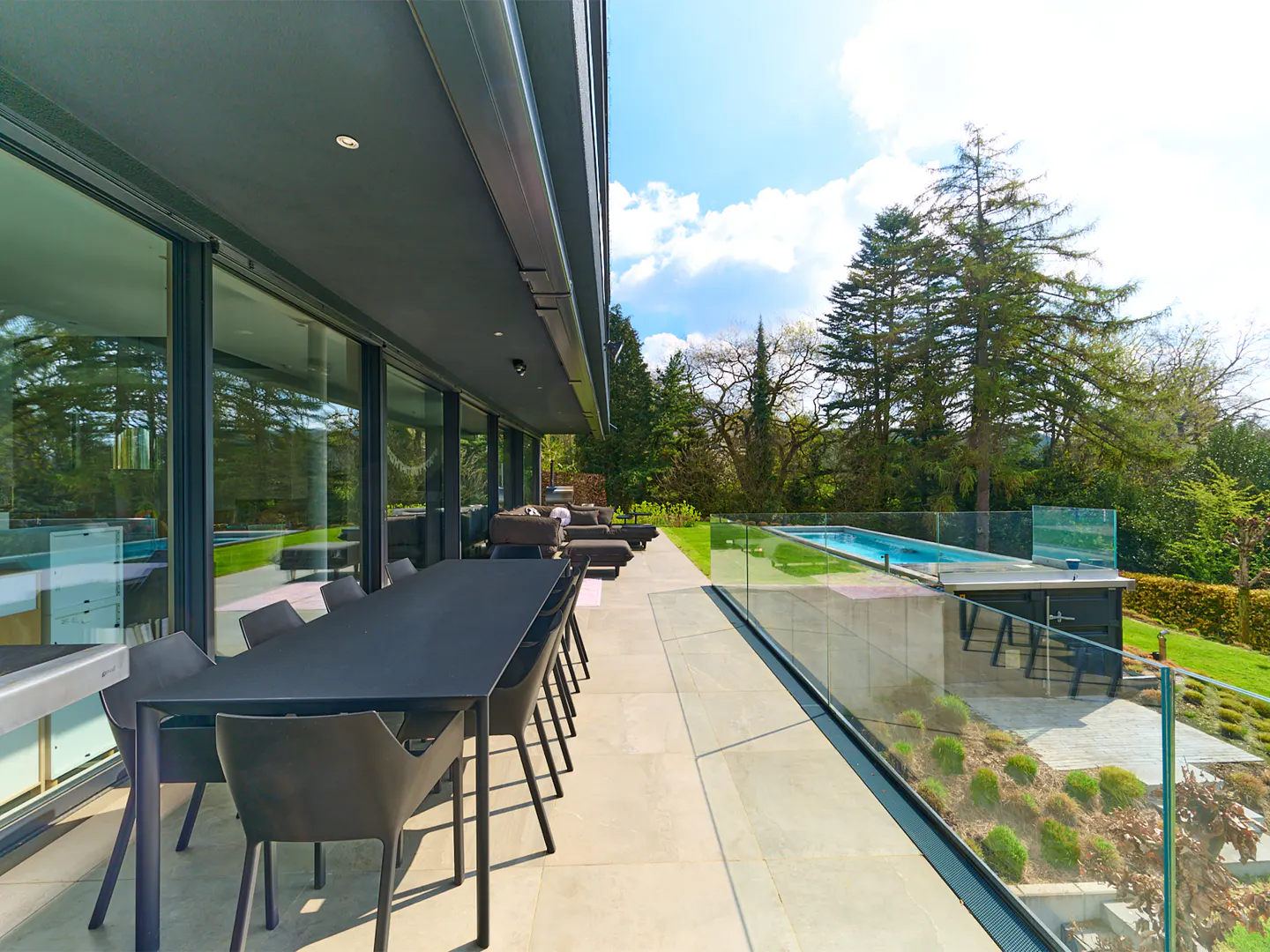 Outdoor patio with a black dining table and chairs, lounge chairs, and a pool with a glass fence.