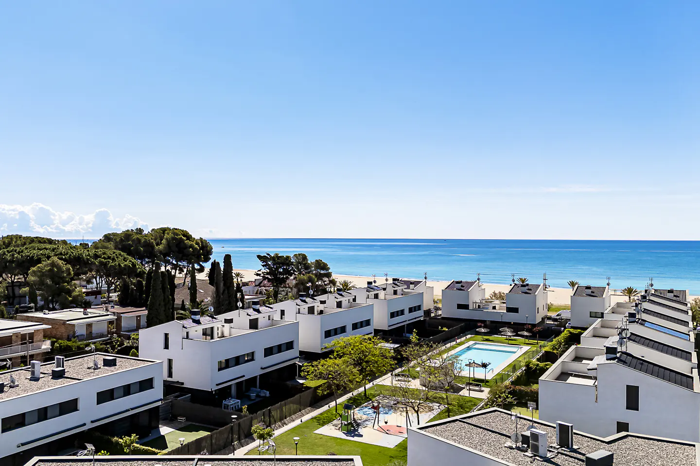 Aerial view of white modern townhouses with a pool, playground, and beach access under a clear blue sky.