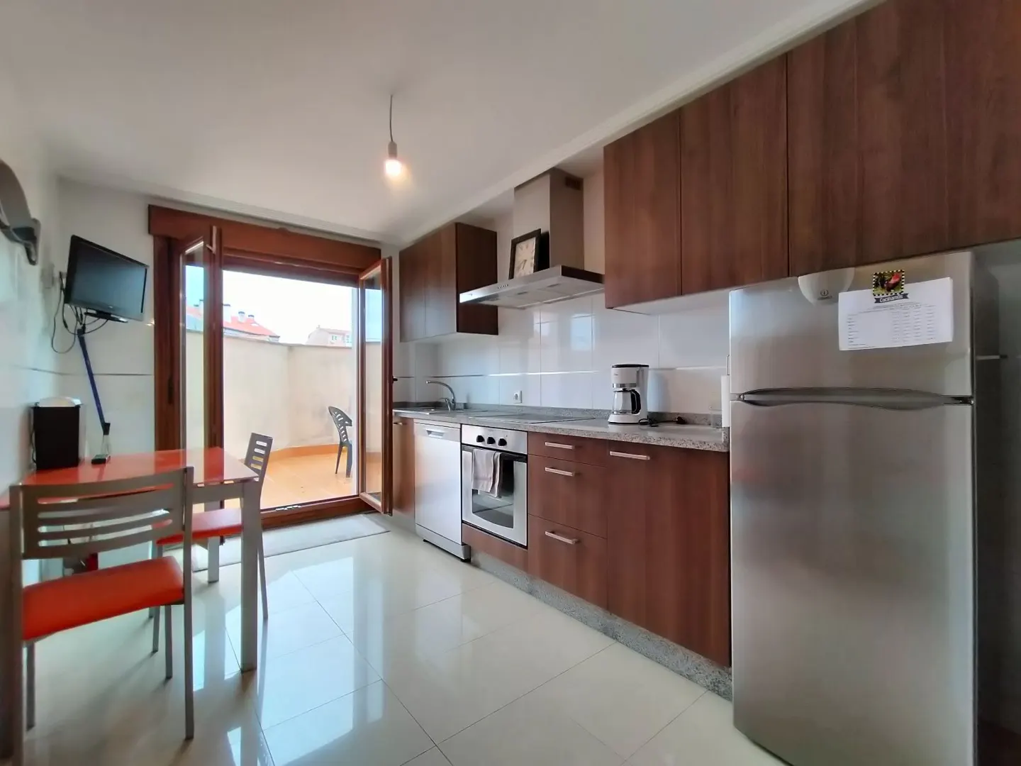 Bright kitchen with brown cabinets, stainless steel appliances, and a dining table with orange chairs. A balcony is visible through open doors.
