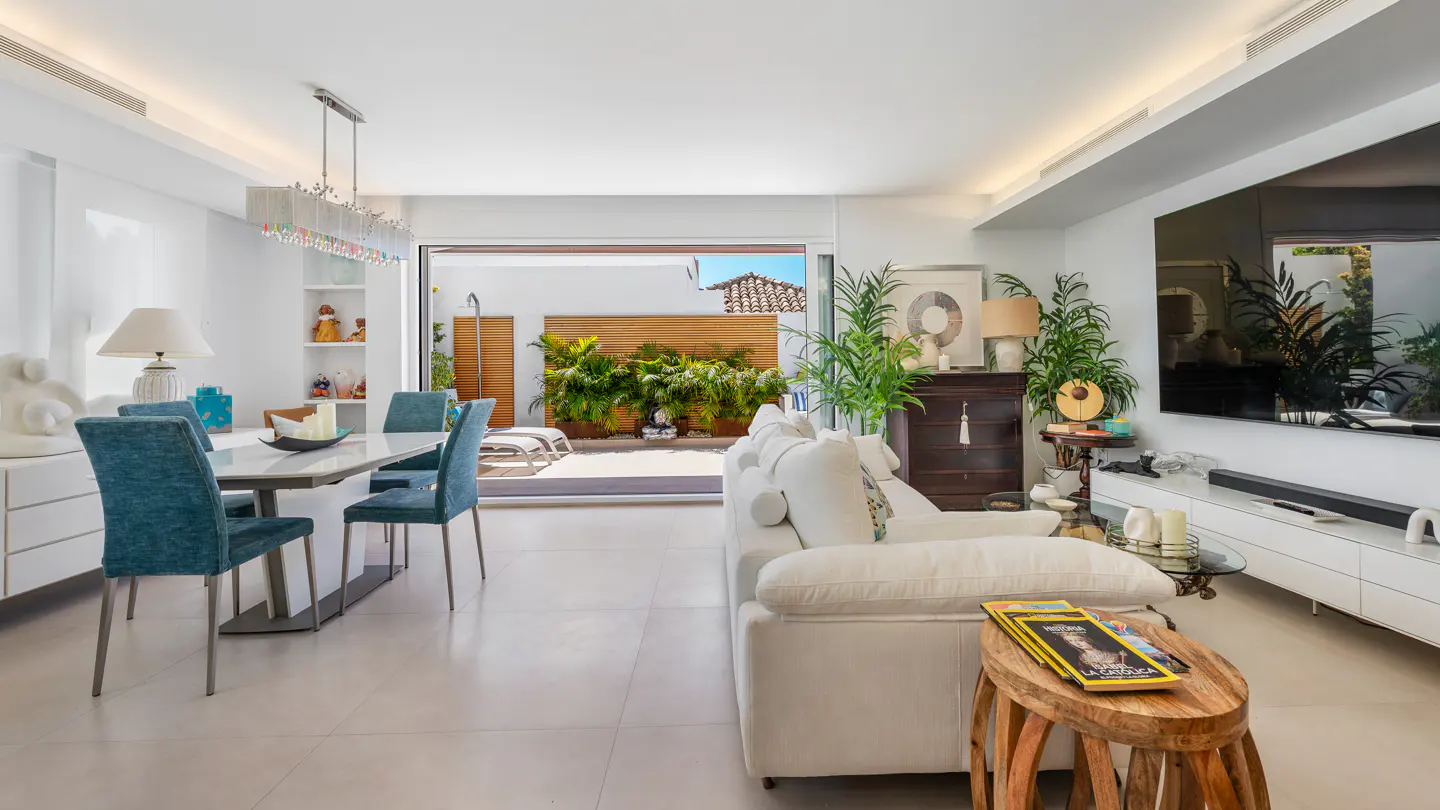 Bright living room with white sofa, teal dining chairs, large TV, and open glass doors leading to a sunny patio garden.