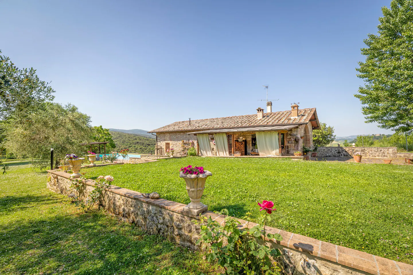 A stone farmhouse with a tiled roof and covered porch sits behind a lush green lawn with a stone wall and flower urns.
