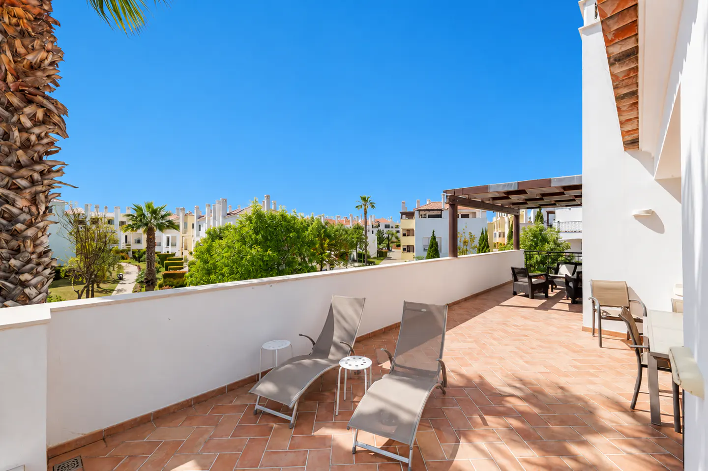 Balcony with two lounge chairs, small tables, and outdoor furniture. View of trees, buildings, and blue sky.