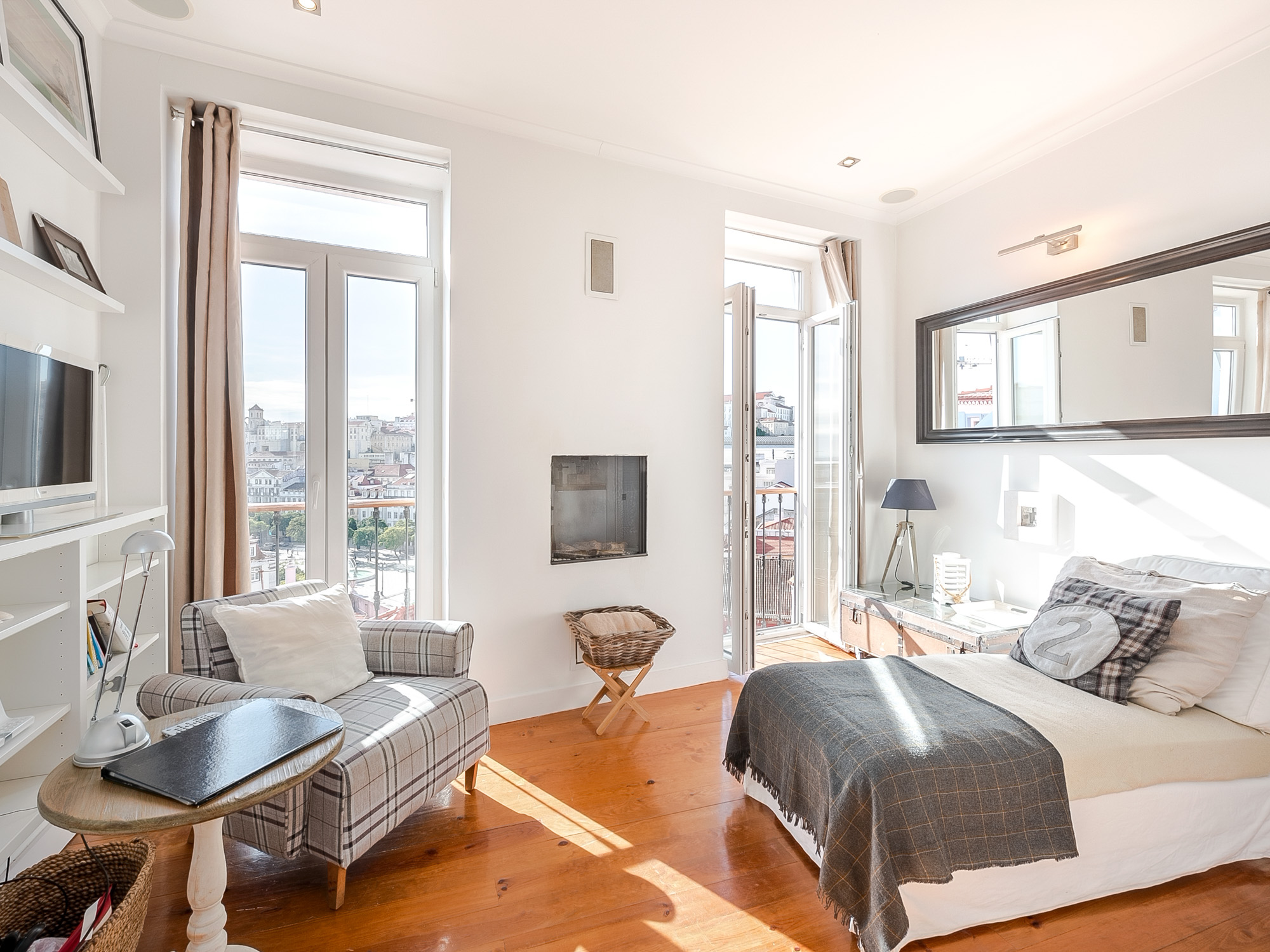Bright bedroom with white walls, wood floors, and city views. Plaid chair, bed with gray blanket, and balcony access.