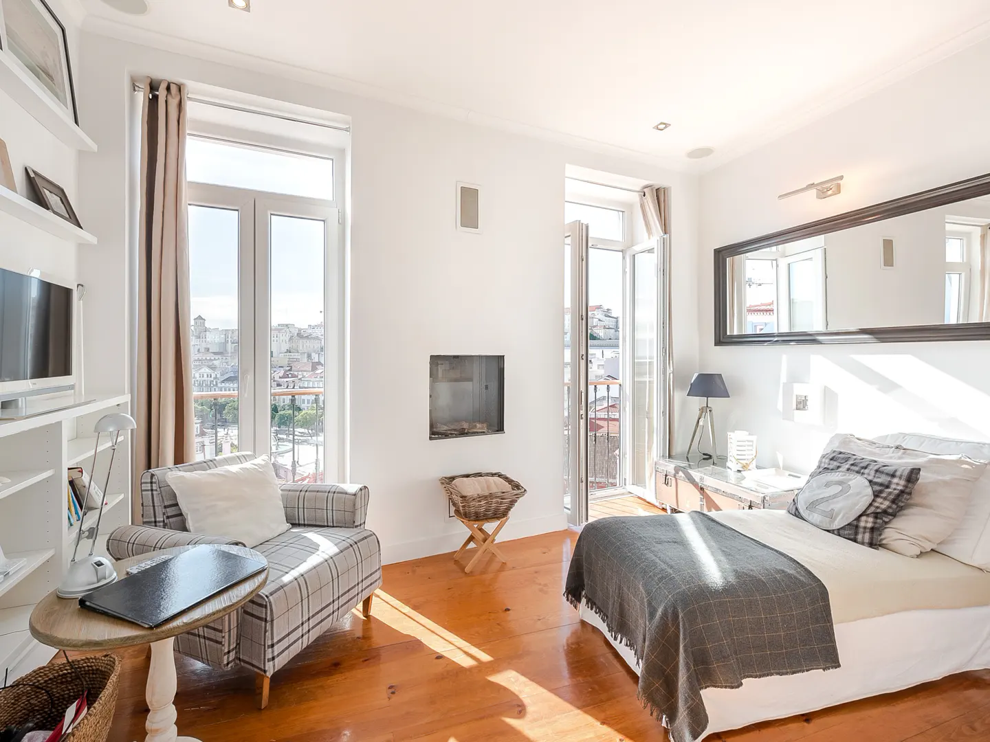 Bright bedroom with white walls, wood floors, and city views. Plaid chair, bed with gray blanket, and balcony access.