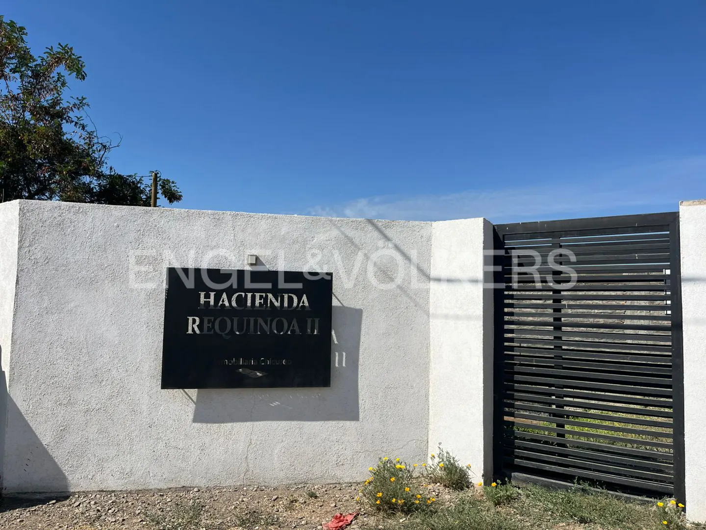 Entrance to Hacienda Requinoa II with a black sign on a white wall and a black metal gate. Blue sky above.