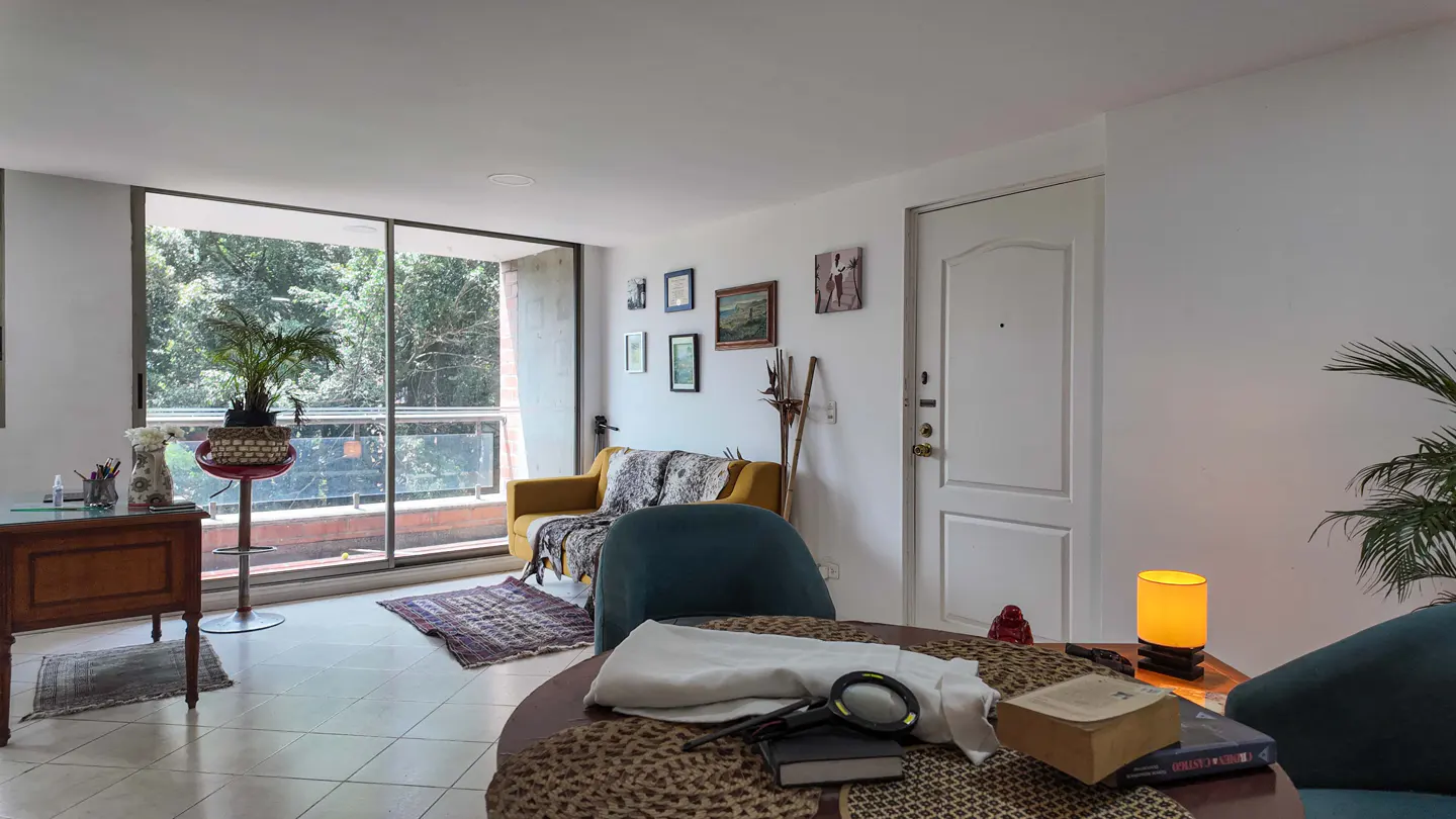 Bright living room with a yellow couch, blue chairs, and a wooden desk by a large window overlooking trees. Artwork adorns the white walls.