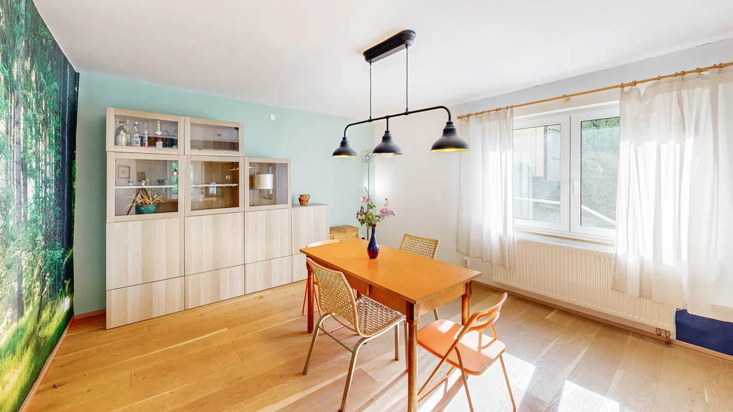 A dining room with a wood table, four chairs, and a black three-bulb light fixture. A forest mural is on the left wall.