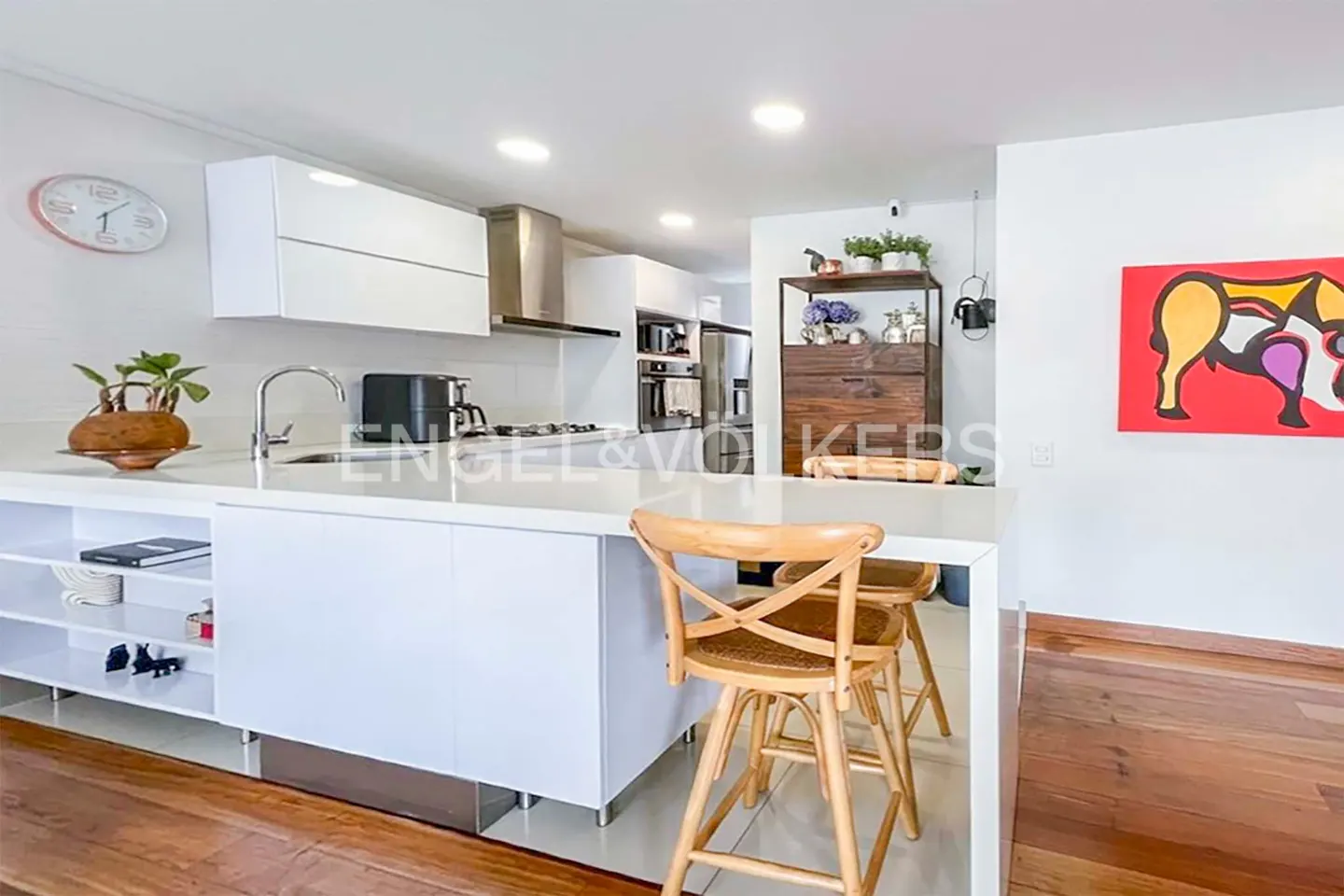 A modern kitchen with white cabinets, stainless steel appliances, and a quartz countertop island with two wooden bar stools. A colorful painting hangs on the wall.