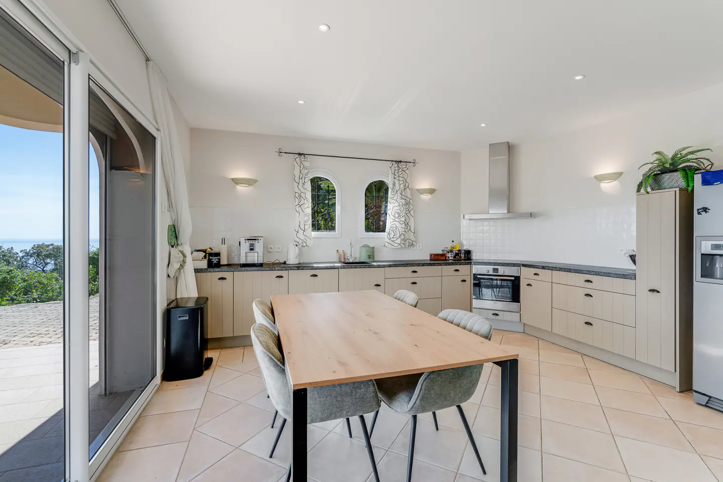 A bright kitchen with beige cabinets, a wooden table with gray chairs, and a view of the ocean through a large sliding glass door.