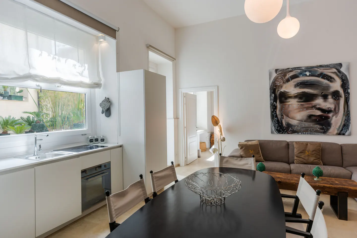 Bright, modern apartment interior with a black dining table, white kitchen, and a large abstract painting above a brown sofa.