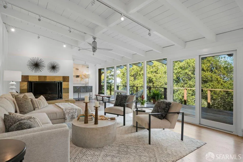 Bright living room with a beige sectional sofa, two armchairs, and a stone coffee table. Large windows overlook a green, wooded area.