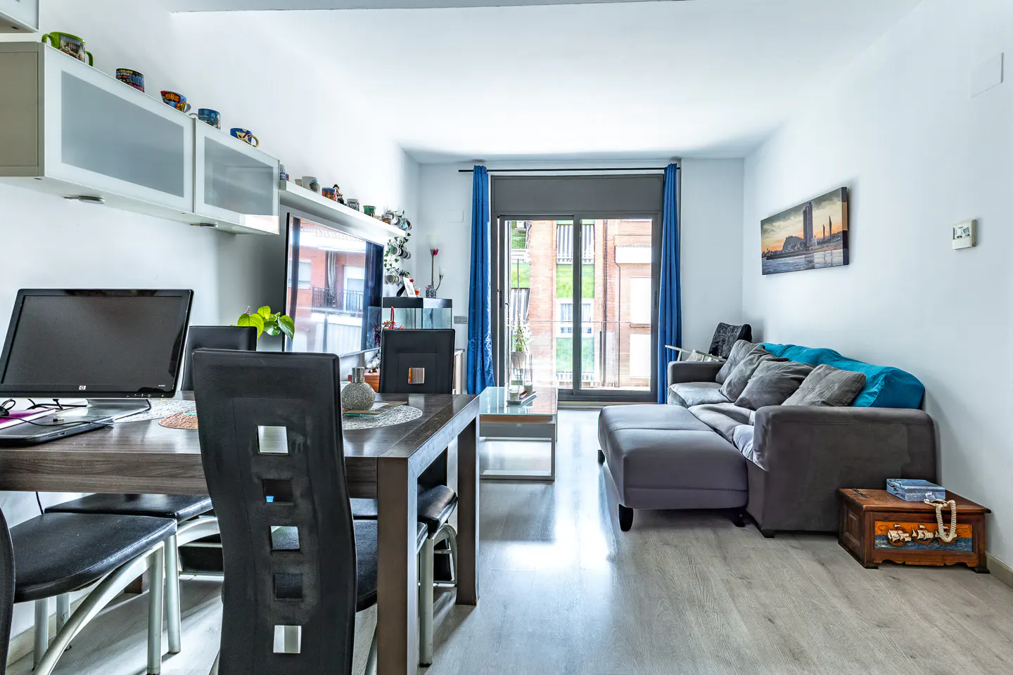 Bright living room with gray sectional sofa, blue accents, and a dining table with a computer. Balcony visible through blue curtains.